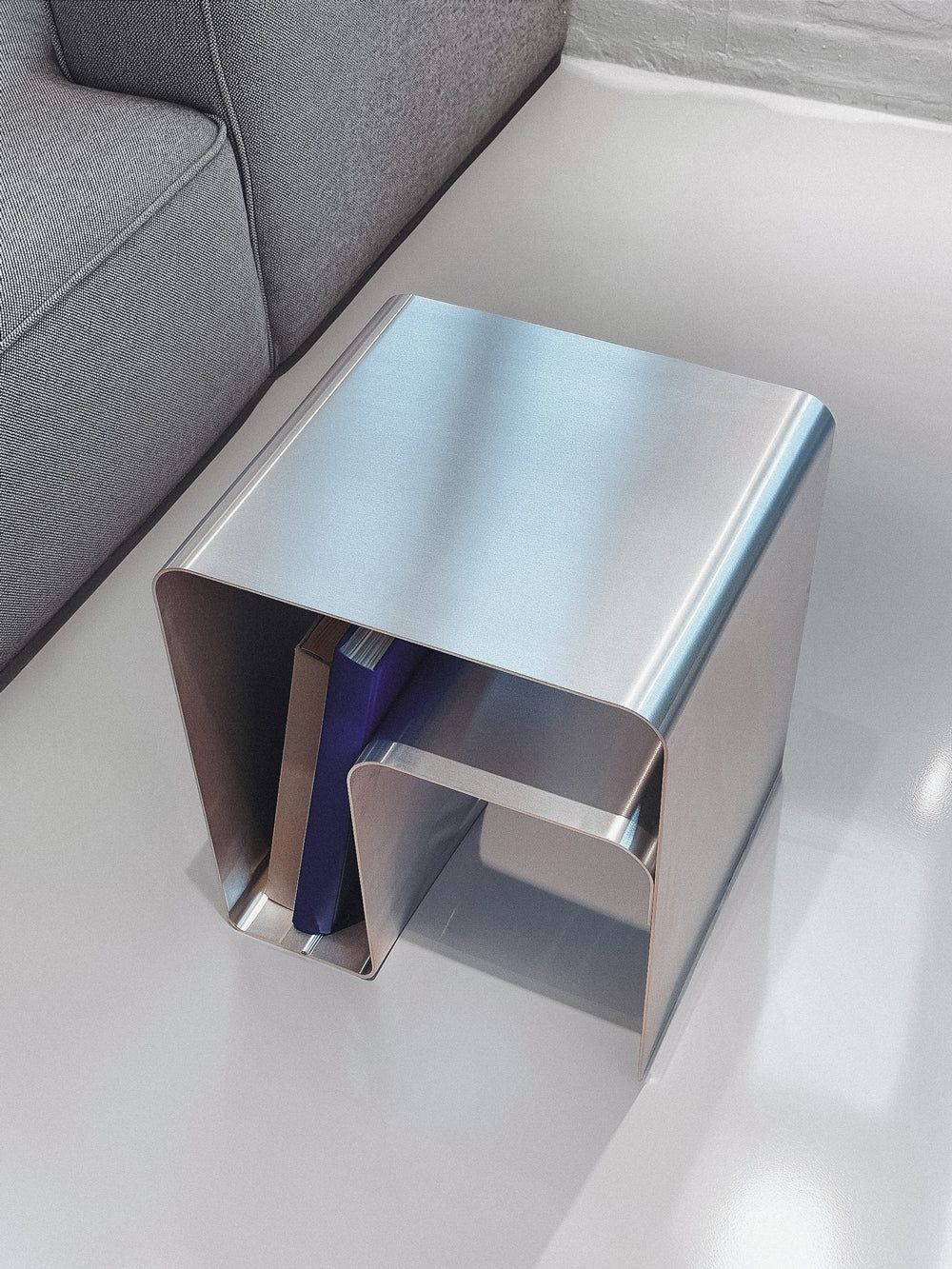 Design sidebord i børstet metal fra Rau Studio med integreret hylde. Sidebord i stål-look med magasinholder.